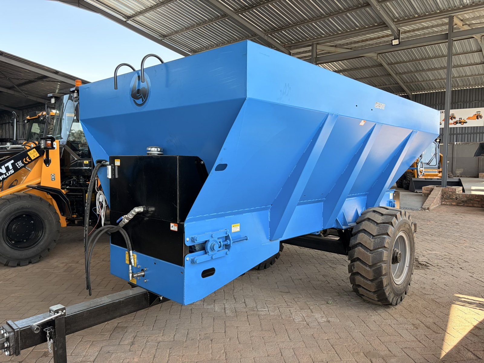 Giant hydraulic manure spreader, front three-quarter view of the hopper, drawbar and single axle, standing under the supplier's canopy
