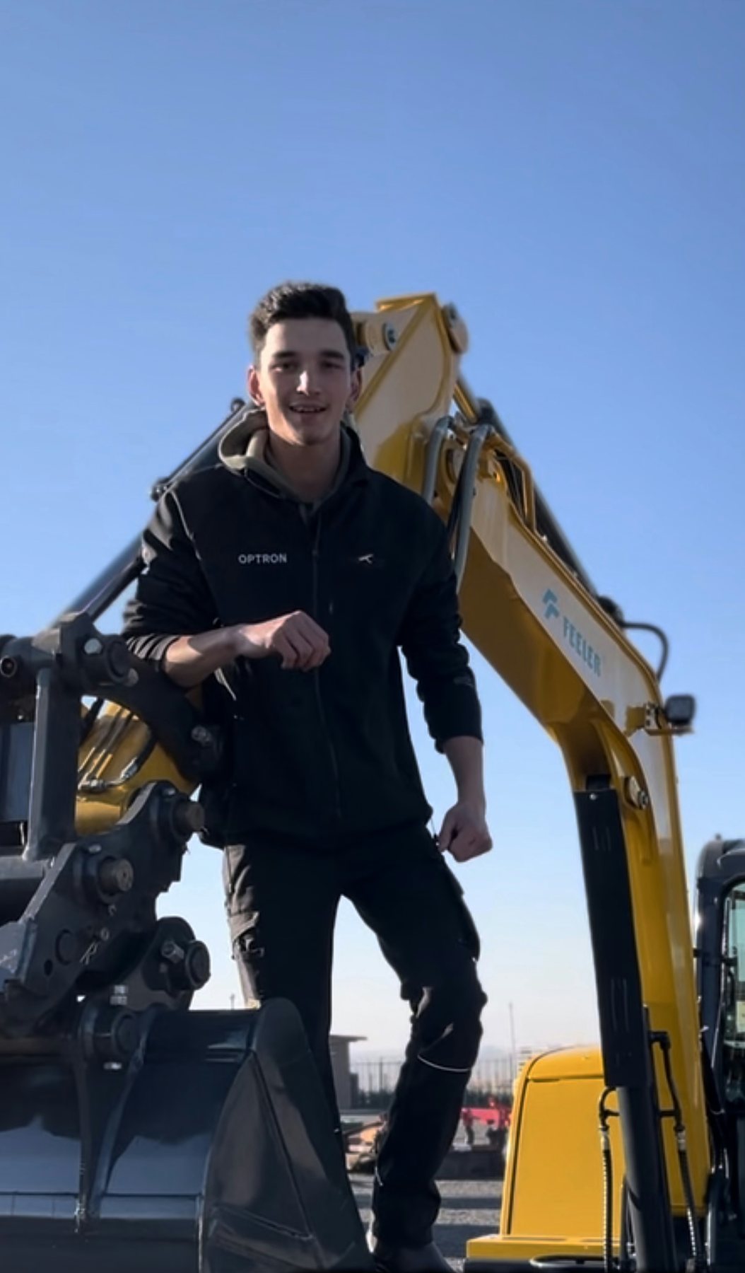 Morne standing on the bucket arm of a yellow excavator at a machinery yard